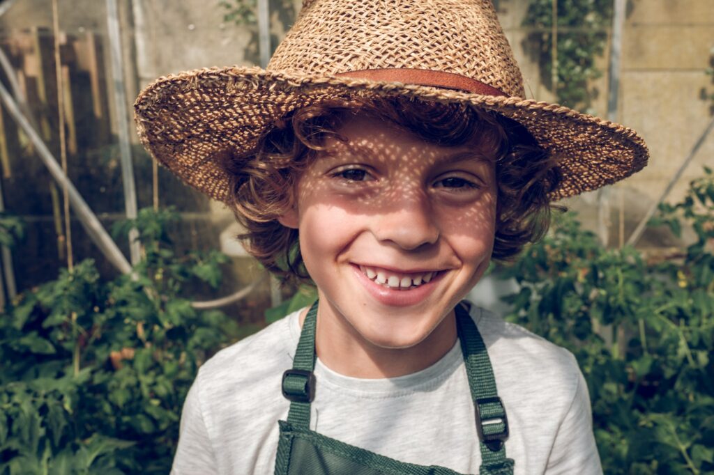 Cheerful preteen boy gardener in wicker hat in greenhouse