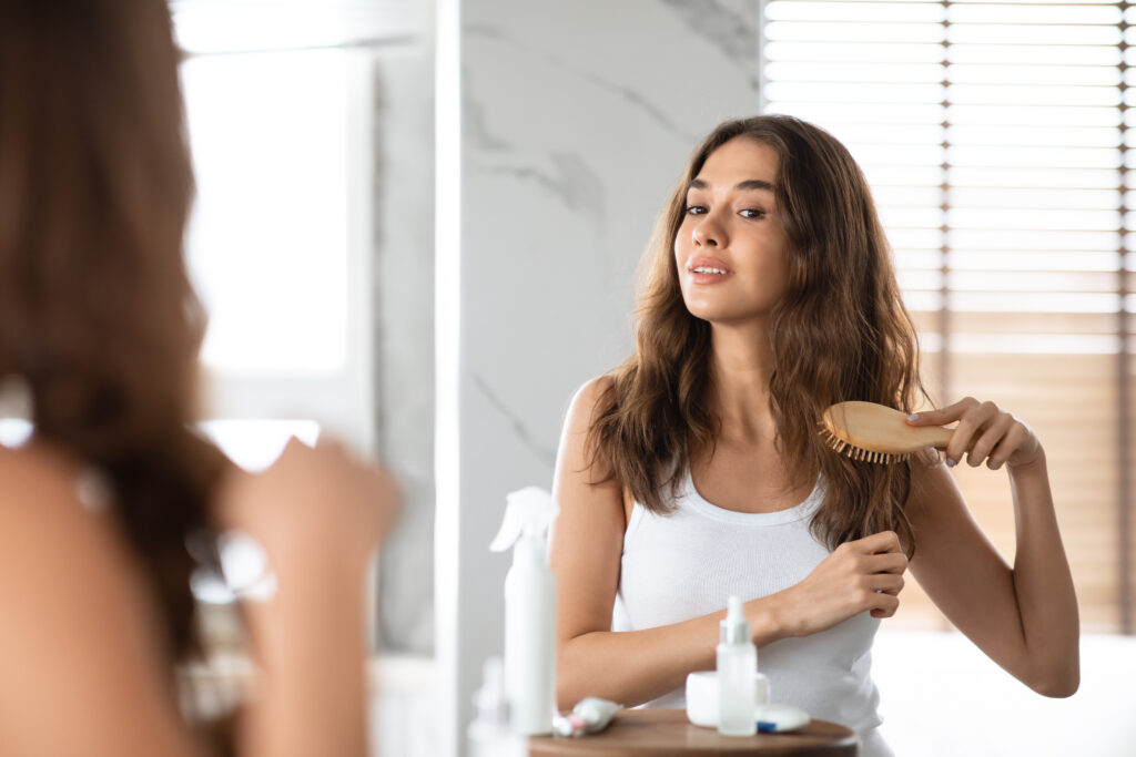 Young Lady Brushing Hair With Hairbrush Standing In Bathroom Indoors