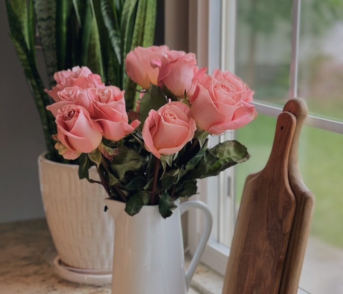A bouquet of soft pink roses in a white pitcher stands next to a window, a potted plant and boards